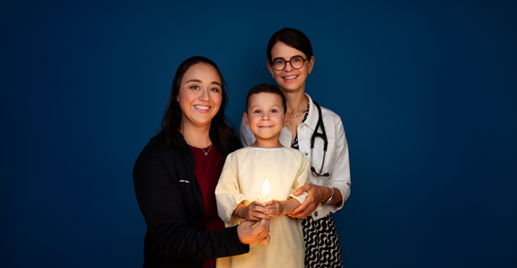 Jacob, 3 ans, porte une jaquette jaune, tient une ampoule allumée dans ses mains, et est entouré de la Dre Sonia Cellot et de son infirmière, Audrey.