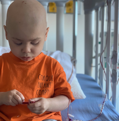 A bald Jacob wearing an orange T-shirt and sitting on his hospital bed.